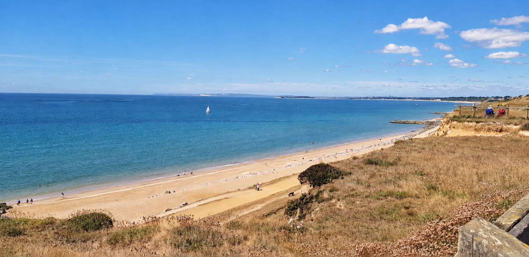 Christchurch Bay on a summer day, with the beach below the cliffs and a sailing boat on the water.