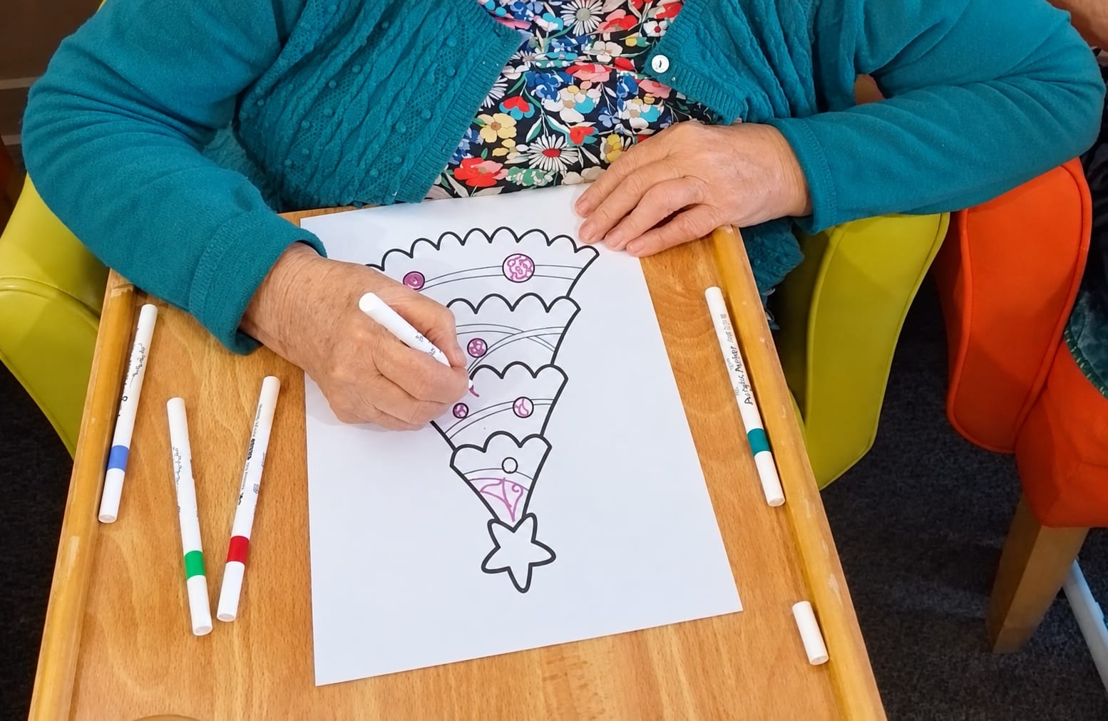 Hands colouring a Christmas tree picture with felt pens on a lap tray.