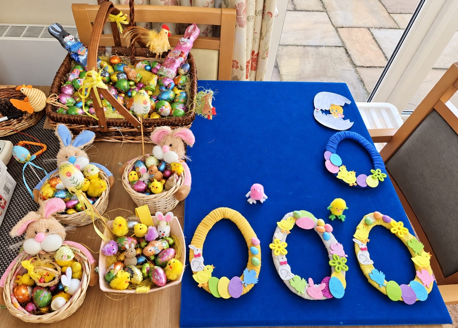 Easter baskets of chocolate eggs, and wreaths with chicks, eggs and flowers, on a blue table.