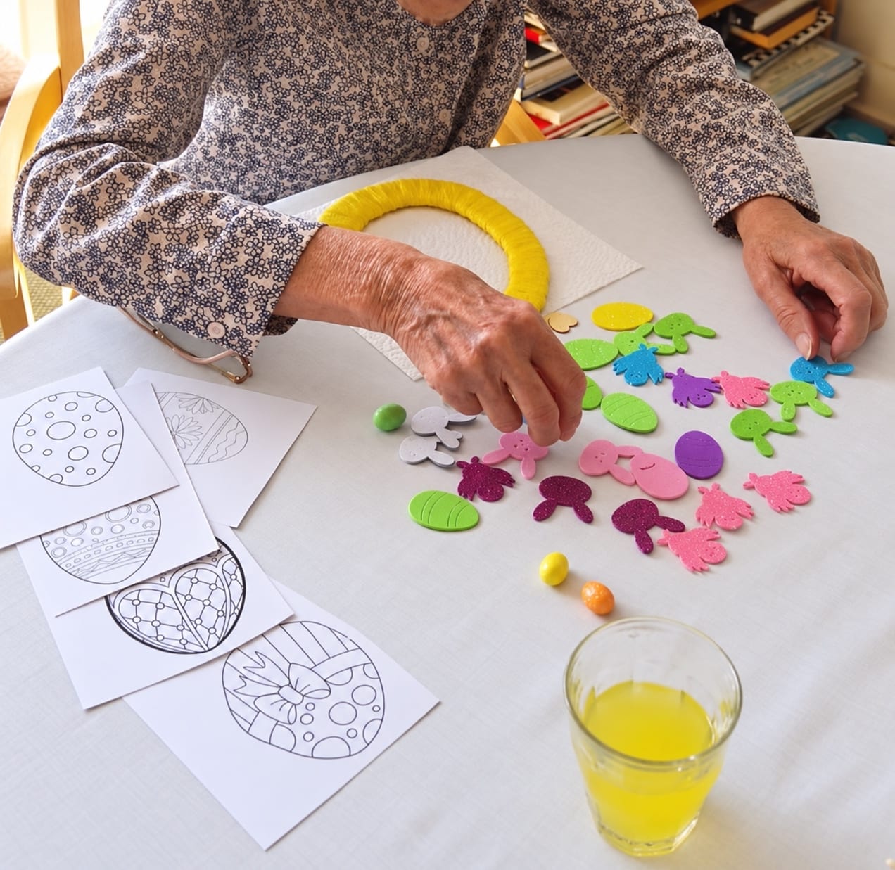 Hands choosing felt eggs and rabbits for a yellow Easter wreath, with colouring sheets on the table.