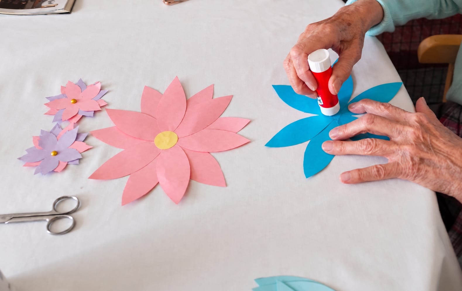 Hands gluing a blue paper flower, with pink paper flowers and scissors on the table.
